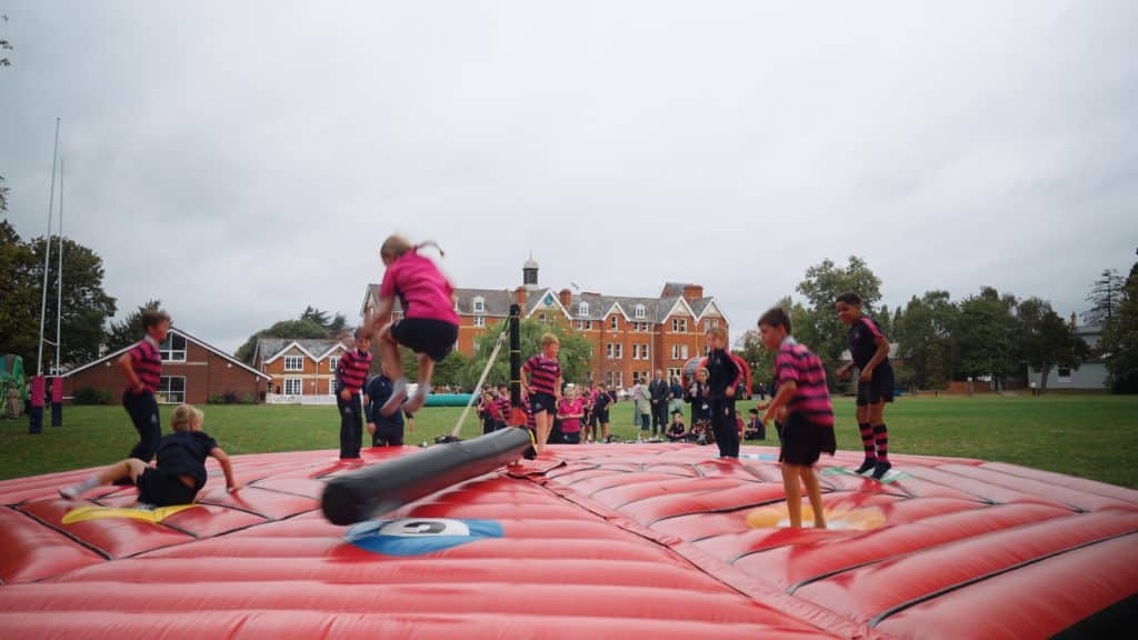Children in striped uniforms play on a large red inflatable game outdoors, jumping and dodging a rotating padded arm, with a brick school building in the background under a cloudy sky.