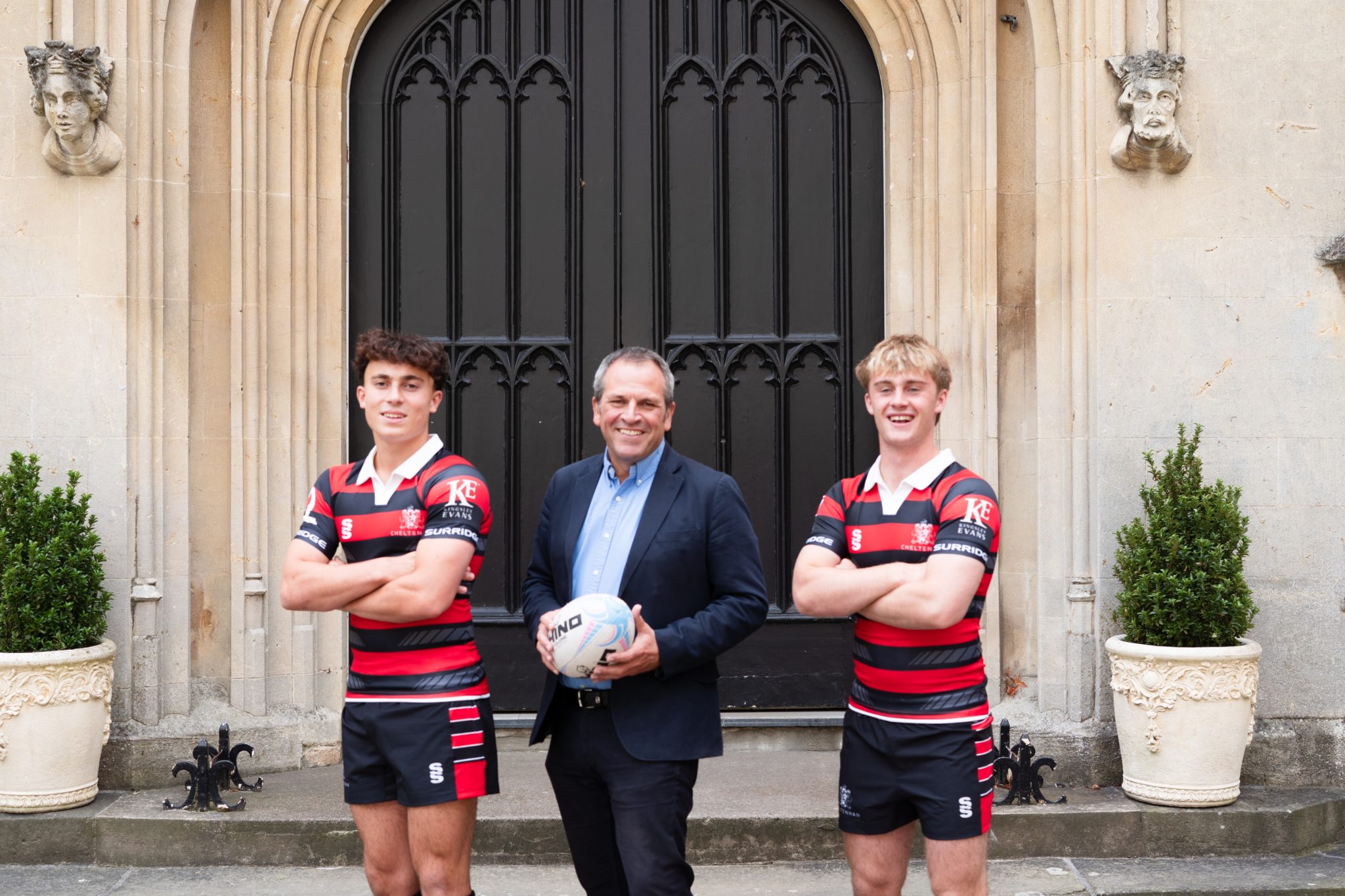 Three men pose in front of a large ornate door; two wear matching black and red striped rugby kits while the man in the centre, dressed in a suit, holds a rugby ball.