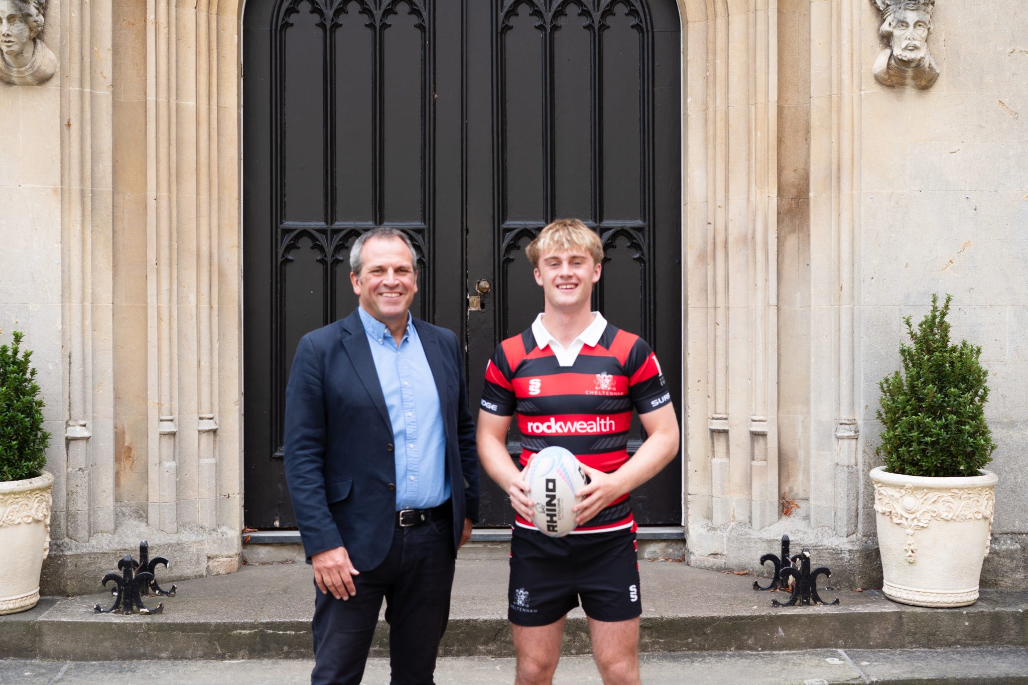 Two men stand outside a stone building; one wears a suit, the other a rugby kit holding a rugby ball. They are smiling, with decorative pots and a black door behind them.