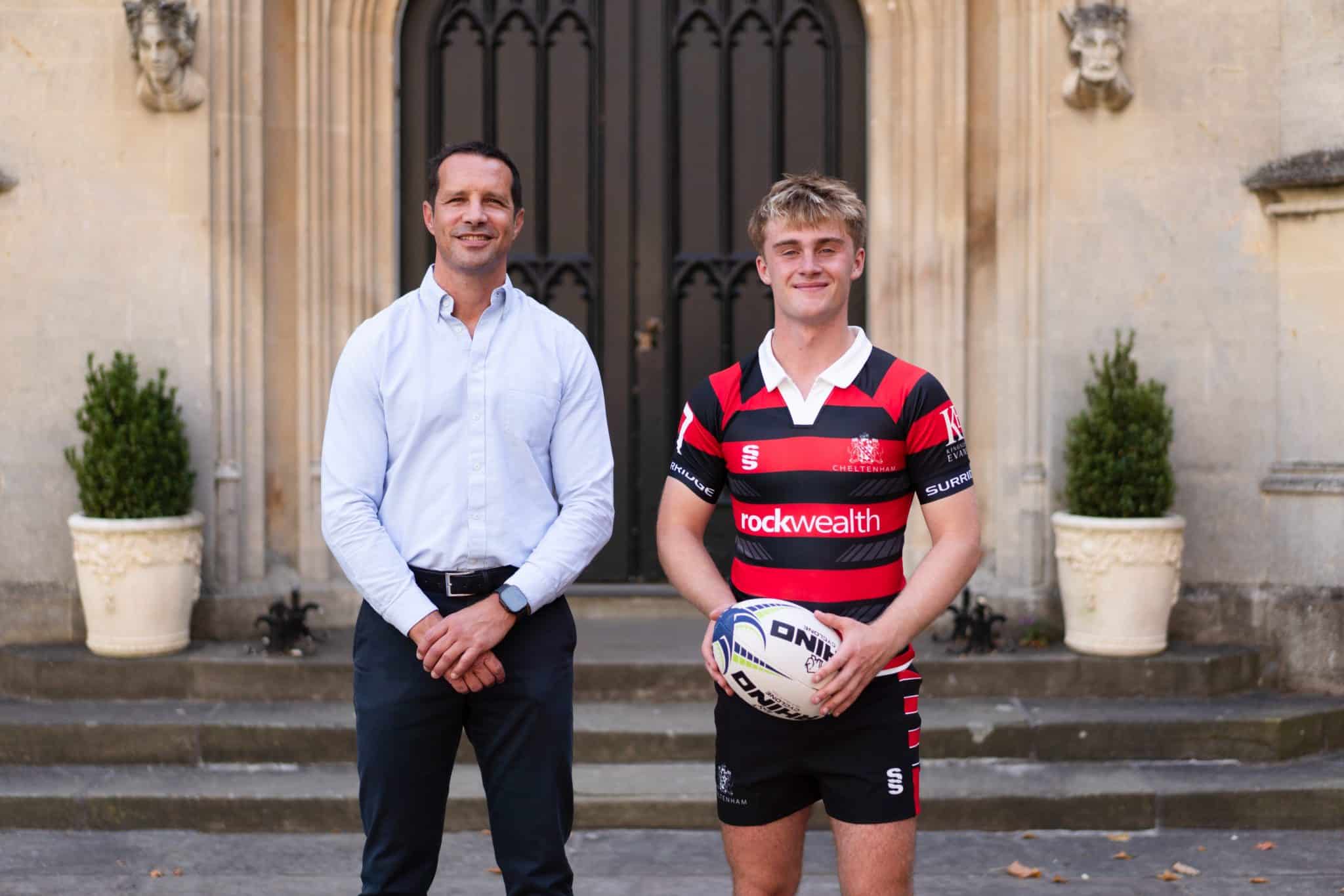 Two men stand in front of an ornate building entrance. One wears a blue shirt and dark trousers, while the other wears a rugby kit and holds a rugby ball. Both are smiling. Potted plants are on either side of the door.