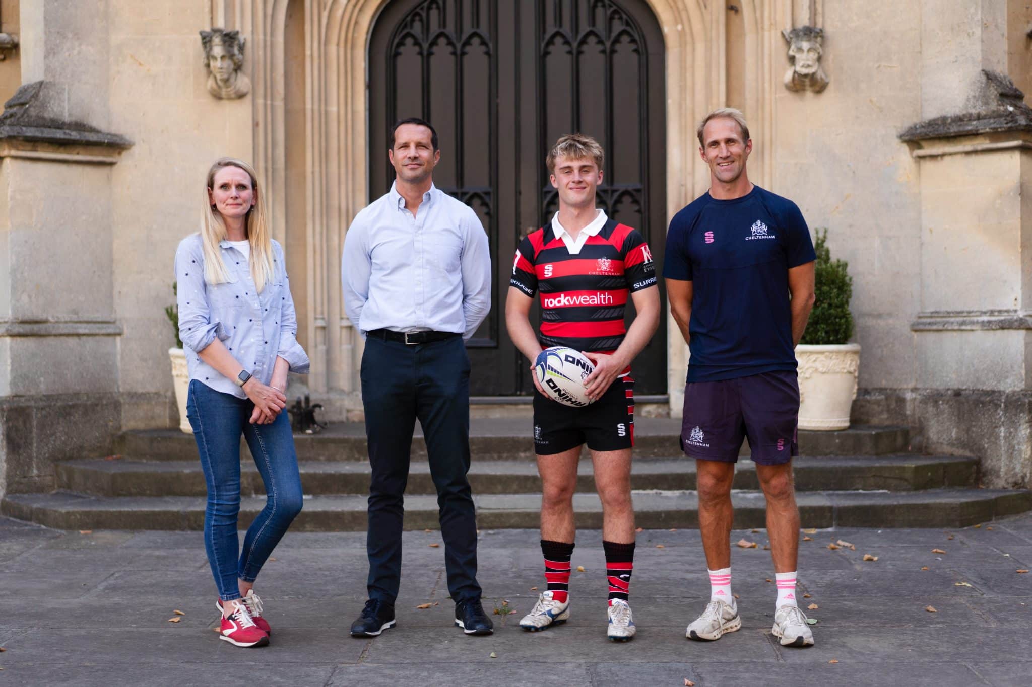 Four people stand in front of an arched doorway; two wear casual clothes, one wears a rugby kit and holds a rugby ball, and one wears athletic gear.