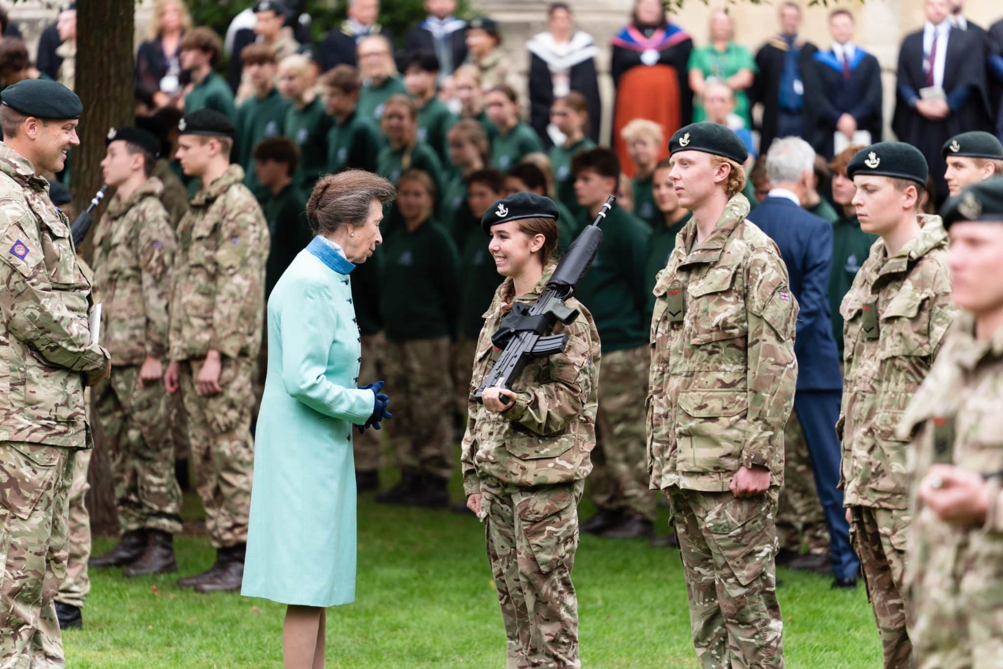 A woman in a light blue coat speaks to a young woman in military uniform holding a rifle. Other soldiers stand nearby in formation, with a large group of people watching in the background.