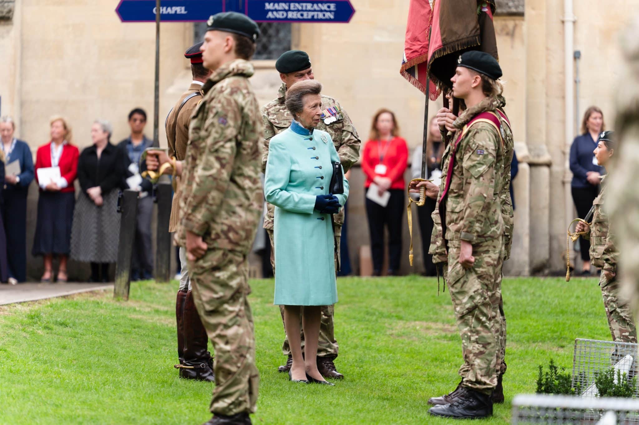 A woman in a light blue coat stands on grass among uniformed soldiers holding flags, with onlookers and a historic building in the background.