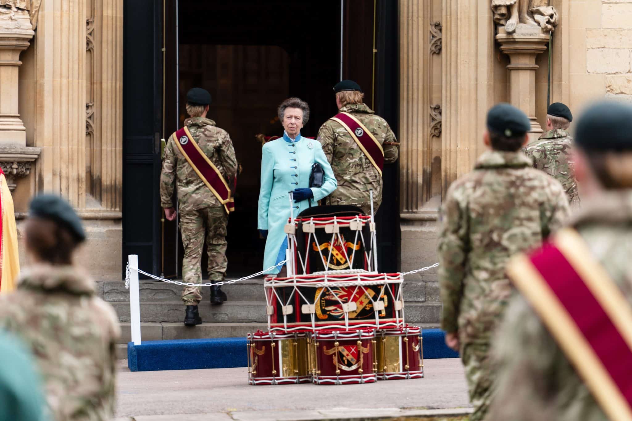 A woman in a light blue coat stands at the entrance of a building, surrounded by soldiers in camouflage uniforms. Drums stacked in front of her, with others marching nearby. Ornate architecture frames the doorway.