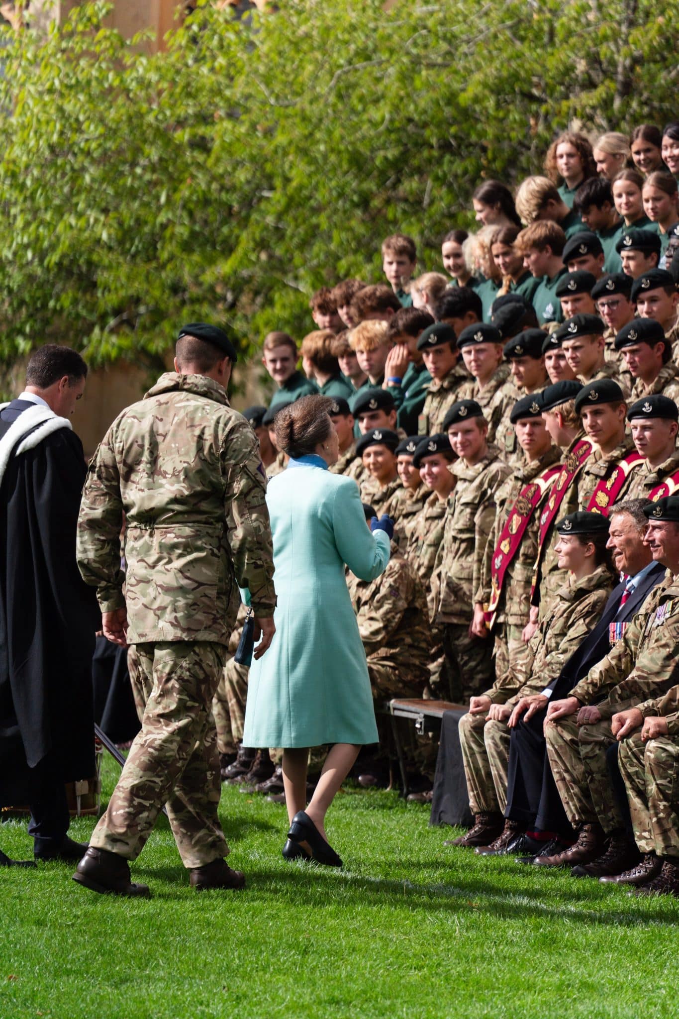 A woman in a light blue coat stands on grass, facing a group of people in military uniforms who are seated and standing outdoors, during a formal event or ceremony.