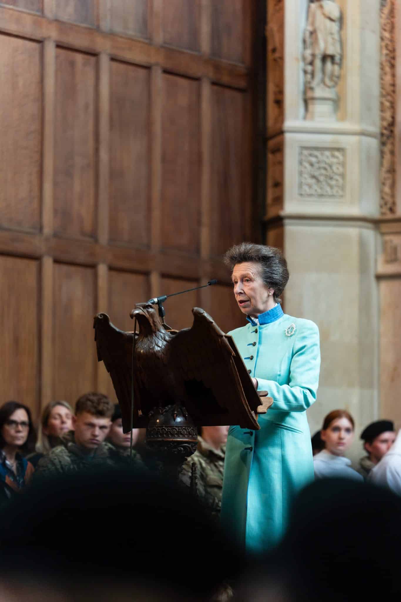 A woman in a light blue dress stands at a podium shaped like an eagle, speaking into a microphone, with an audience seated behind her in a wood-panelled room.