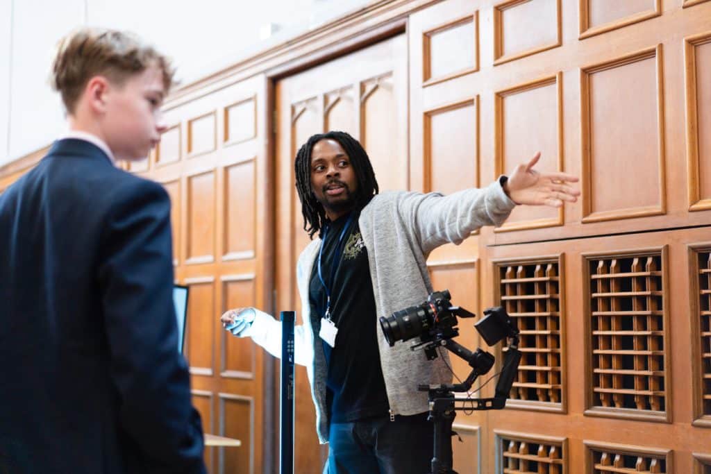 A man gestures and speaks to a boy in a suit near a camera set-up in a room with wooden panelling. The man appears to be explaining something, and the boy listens attentively.