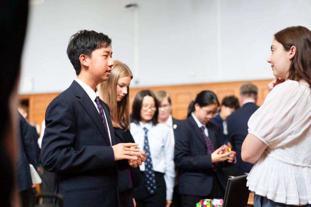 A group of students in uniforms stand and talk indoors, some holding Rubik's cubes, whilst a woman in a white blouse listens to a boy in a suit.