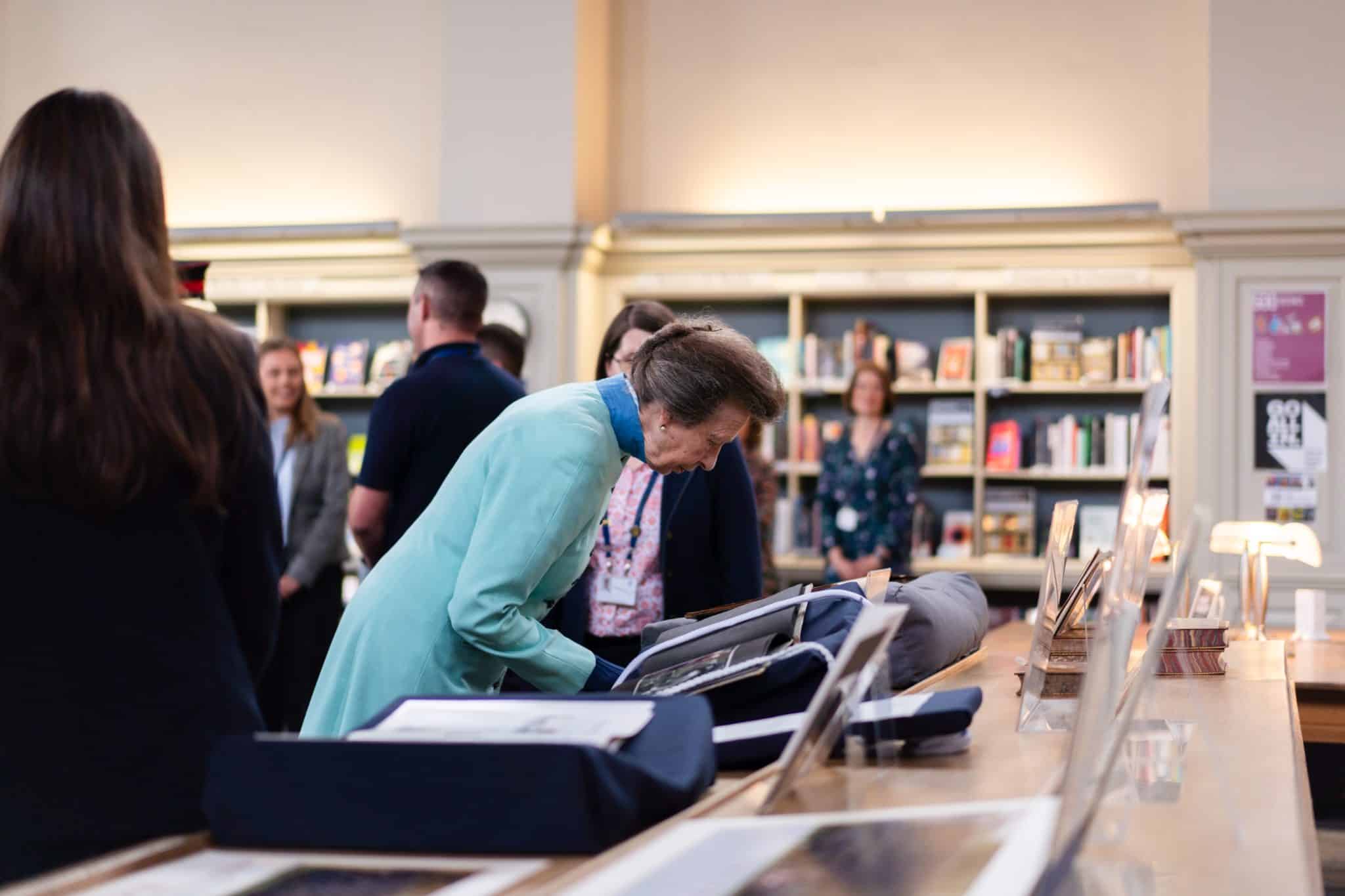 A woman in a light blue coat bends over to look at open books and documents on display in a library, whilst several people stand and observe in the background.