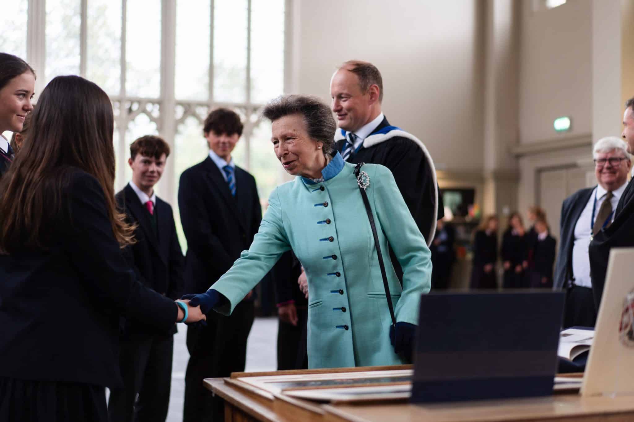 A woman in a light blue coat shakes hands with a student in a group of people dressed in formal attire inside a bright, elegant room with large windows.
