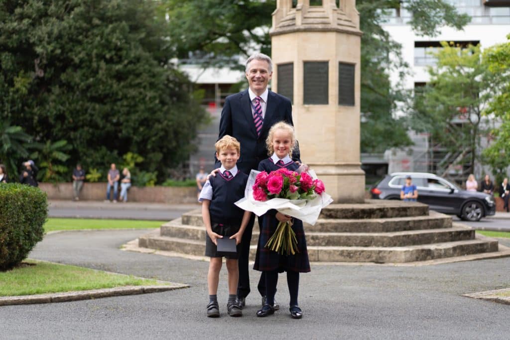 A man in a suit stands behind two children in school uniforms. The girl holds a large bouquet of pink flowers, and they all smile, posing in front of a stone monument in a park—a memorable royal occasion for prep pupils.