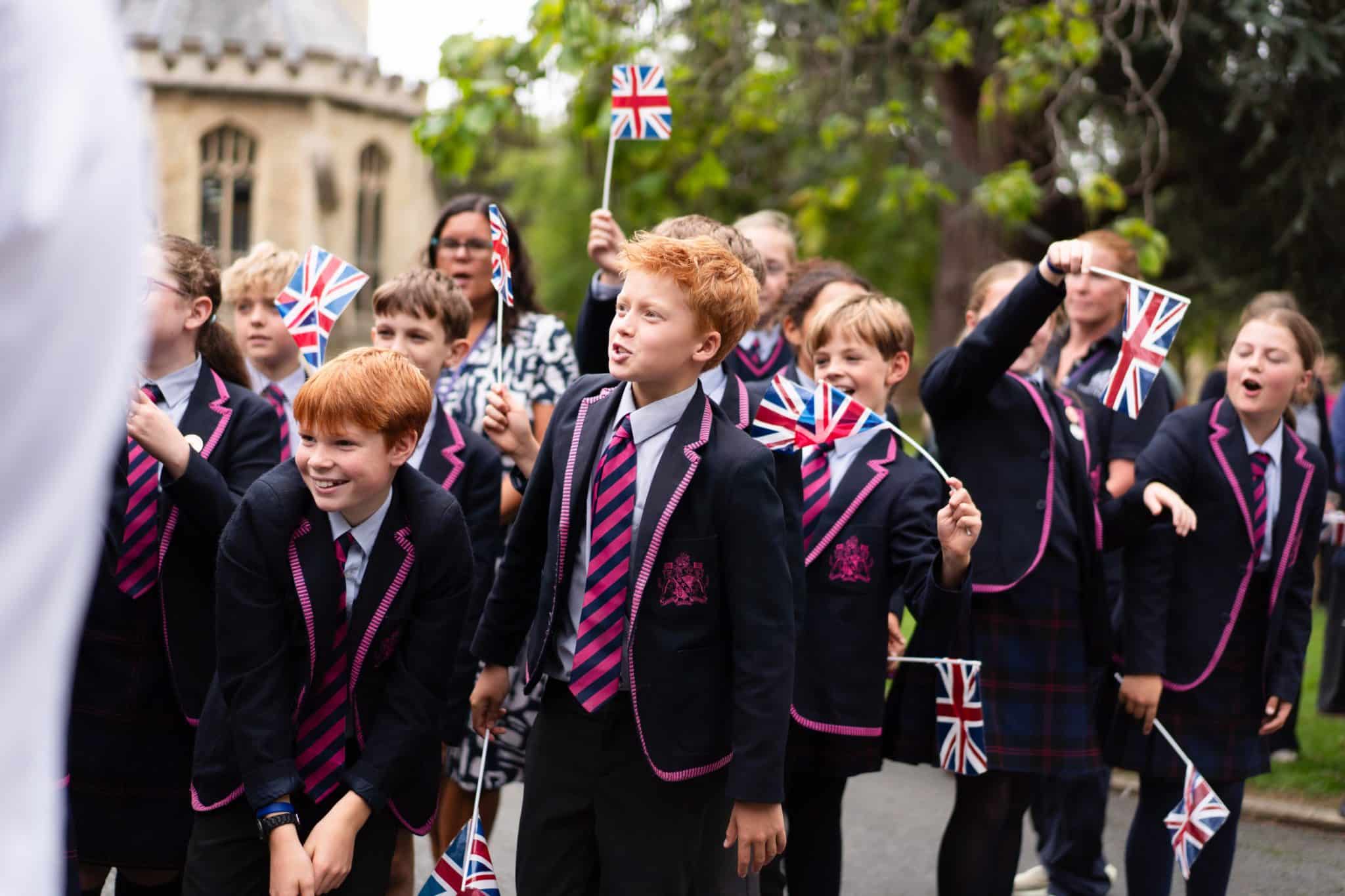 A group of schoolchildren in uniforms wave small UK flags outdoors, smiling and excited. Some are singing or shouting. Trees and a building are visible in the background.