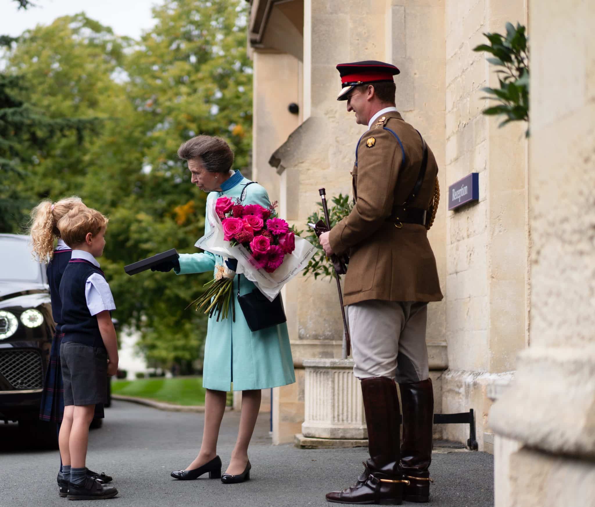 A woman in a light blue coat holding flowers greets a young girl in uniform outside a building, while a man in a brown military uniform stands nearby, smiling.