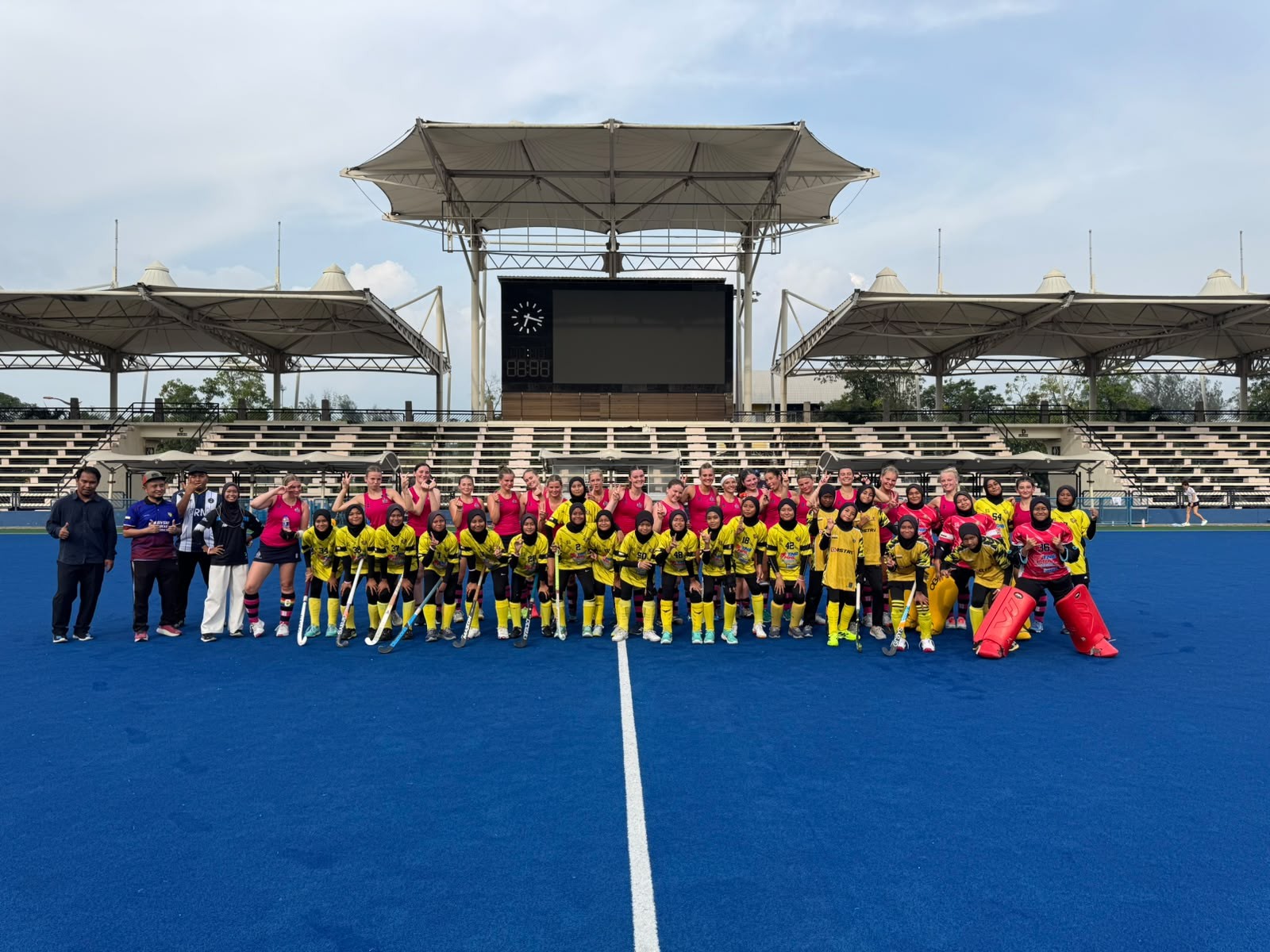 A group of field hockey players in red and yellow kits pose together on a blue hockey pitch, with coaches and staff, in front of stadium seats and a canopy roof.