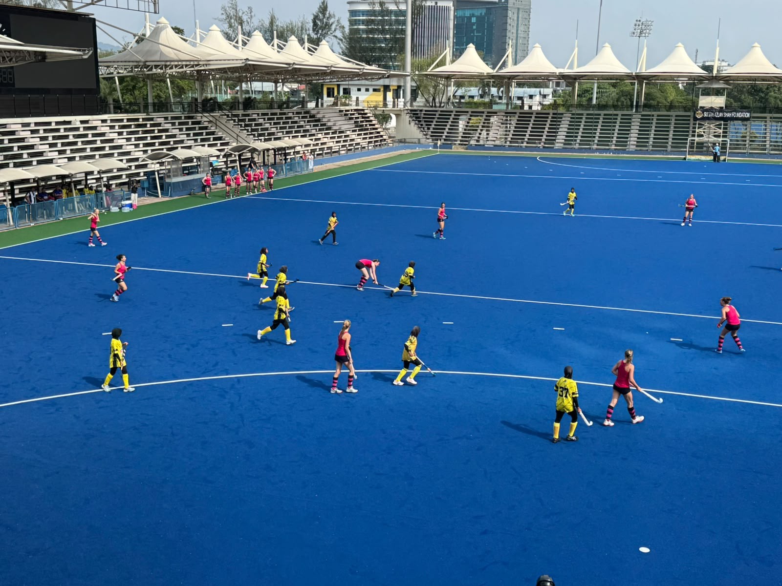 A field hockey match on a blue pitch shows players in yellow and black kits facing off against players in red and white kits, with empty stands and a modern building in the background.