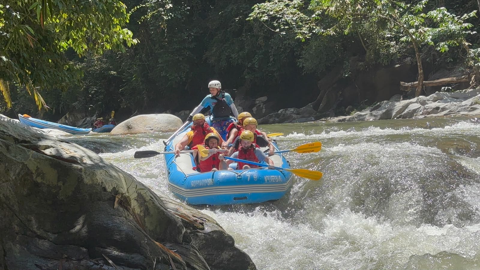 A group of people wearing helmets and life jackets are white-water rafting in a blue inflatable boat through a rocky river surrounded by lush green trees.