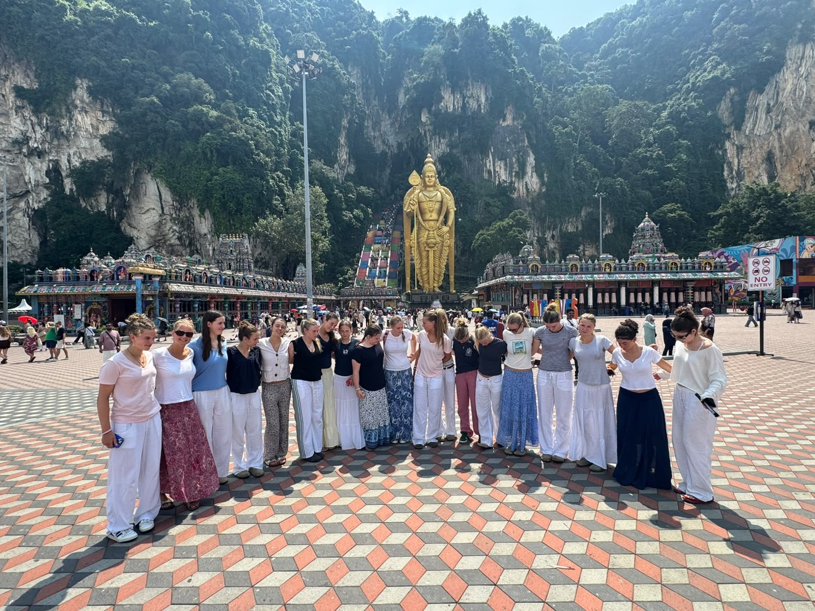 A group of people stands in a semi-circle at Batu Caves in Malaysia, with a large golden statue and colourful temple steps visible in the background under a sunny sky.