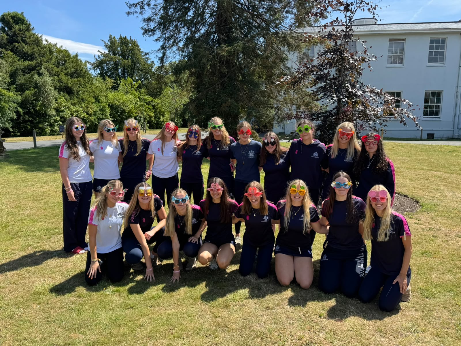 A group of young women wearing colourful novelty glasses pose and smile together on a grassy lawn in front of a white building and trees, enjoying a sunny day.