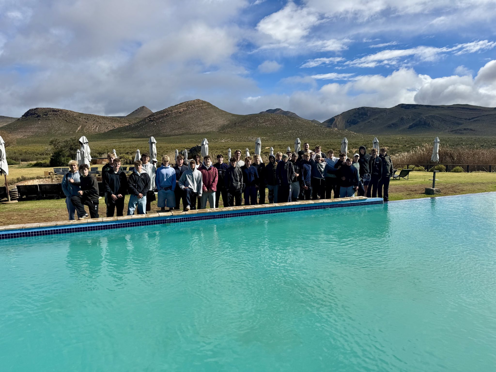 A large group of people stand together beside a clear blue swimming pool, with green hills and mountains in the background under a partly cloudy sky.
