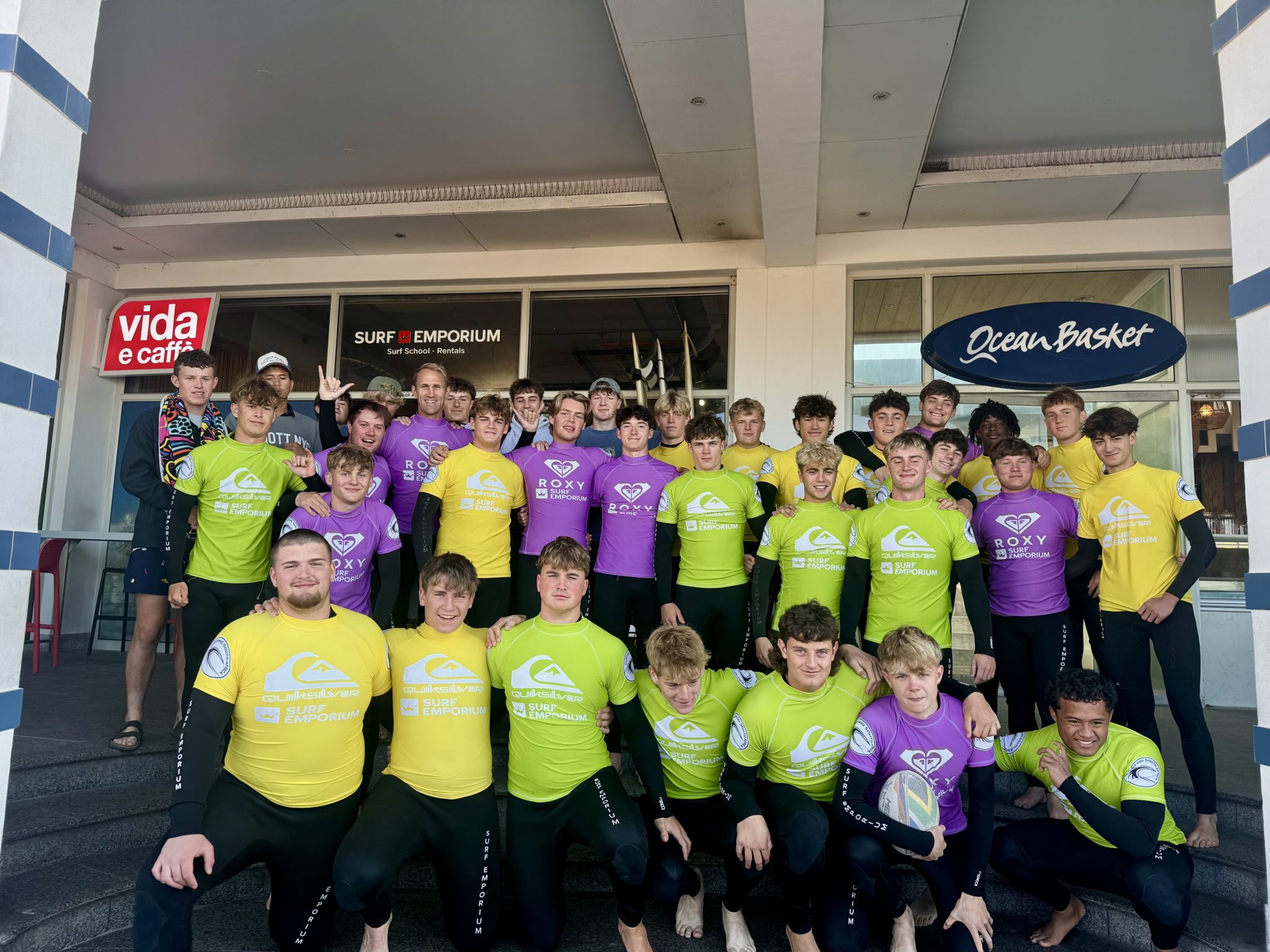 A large group of young people wearing yellow and purple wetsuits pose for a photo on the steps outside surf shops, with Surf Emporium and Ocean Basket signs visible behind them.