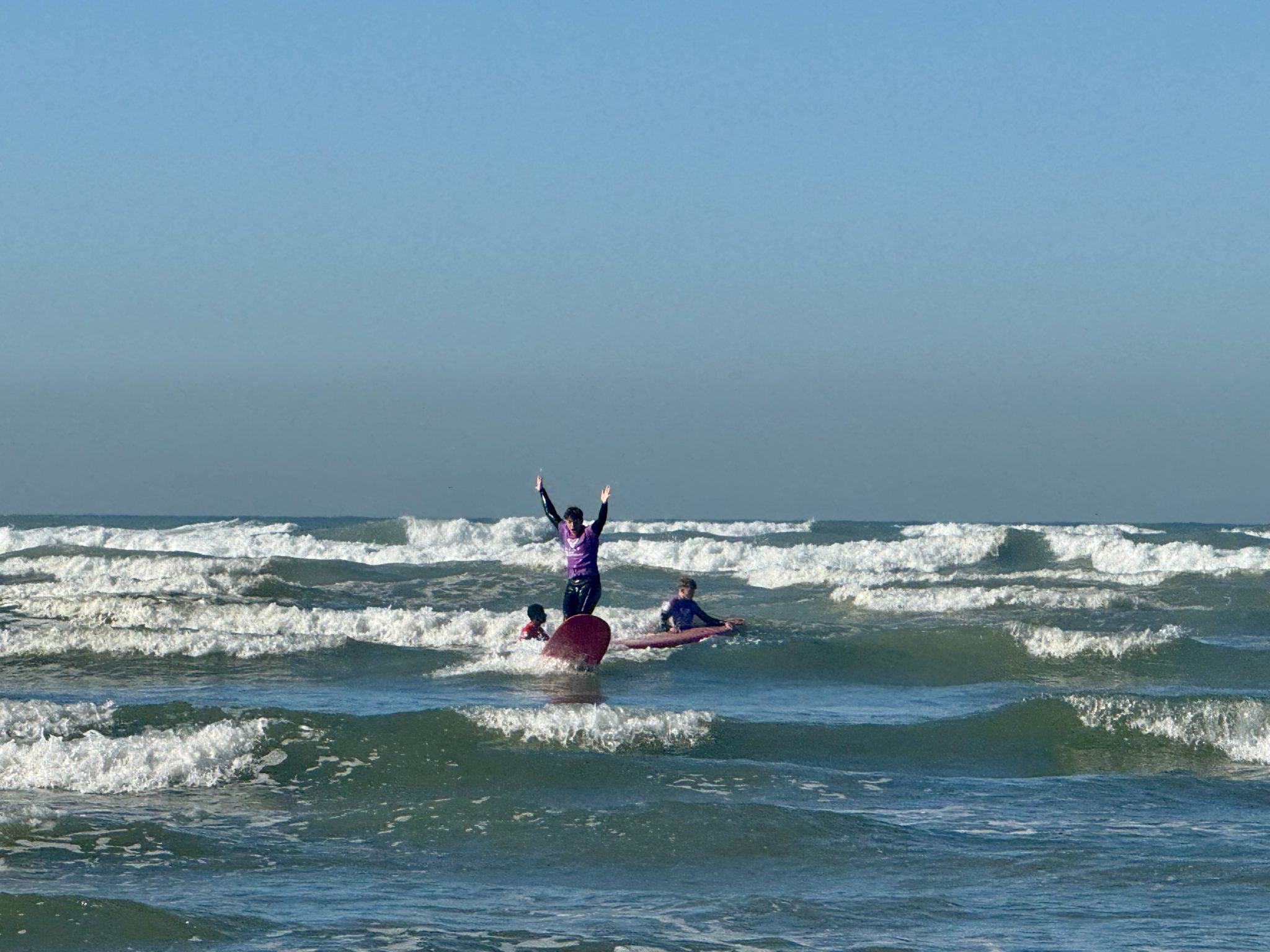 Three people are surfing in the sea with choppy waves. One person stands on a surfboard with arms raised in triumph whilst the others paddle nearby. The sky is clear and blue.