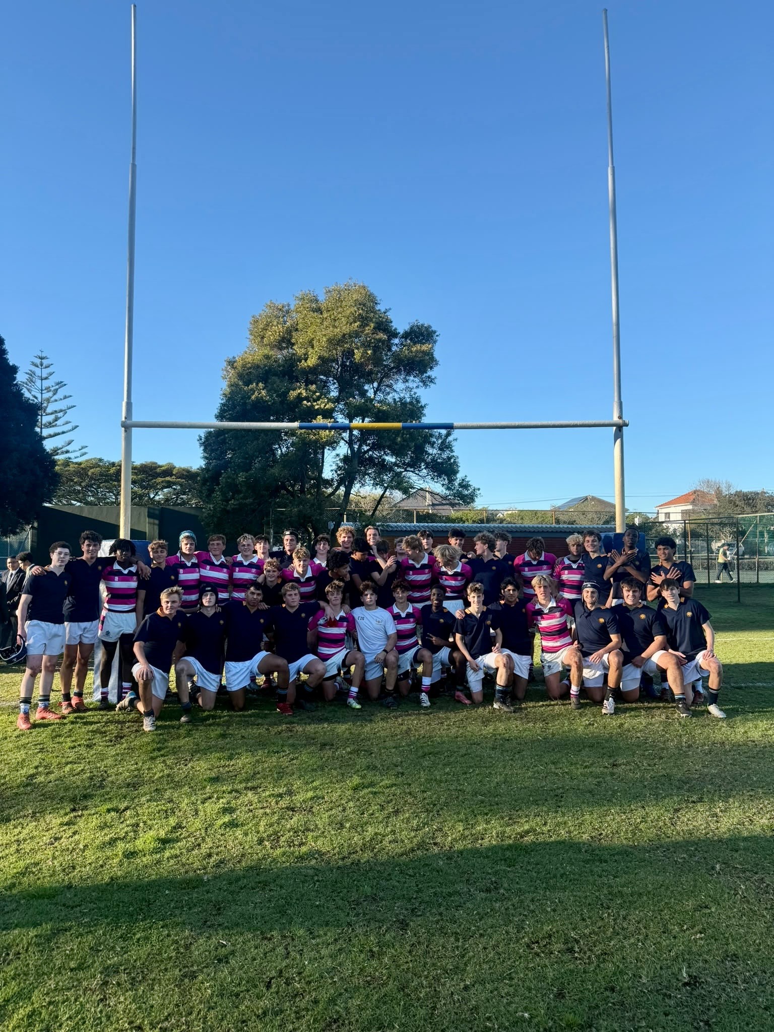 A group of rugby players pose together under goalposts on a grassy pitch, some wearing striped jerseys and others in dark shirts, with trees and houses in the background.