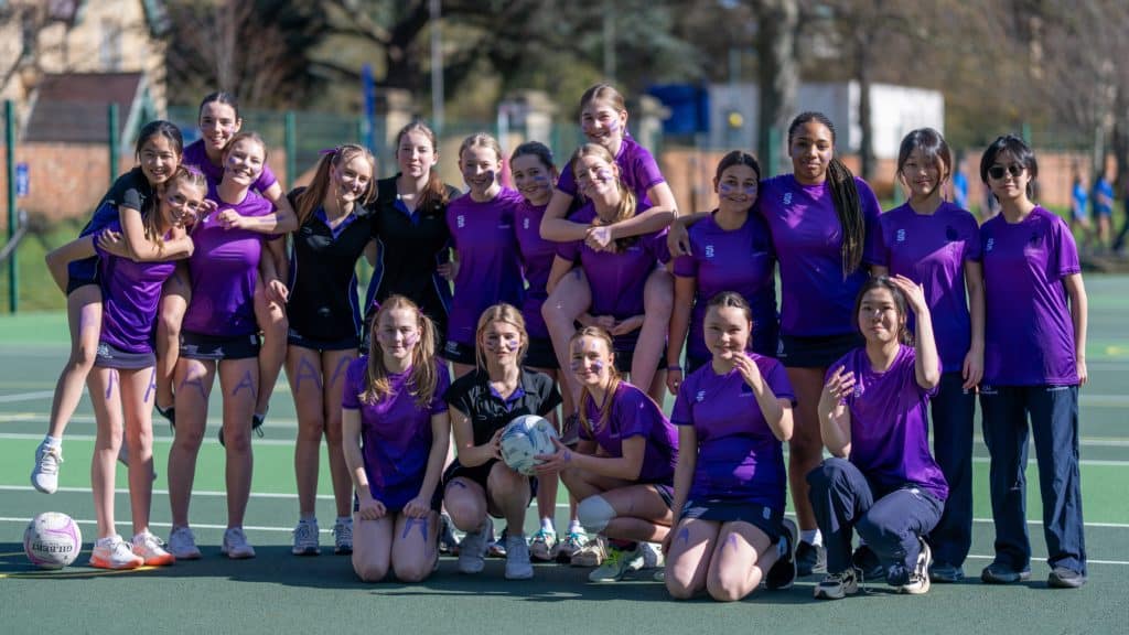 A large group of Cheltenham College Houses students in matching purple sports kits posing on a sports court, with one student in the centre holding a netball.