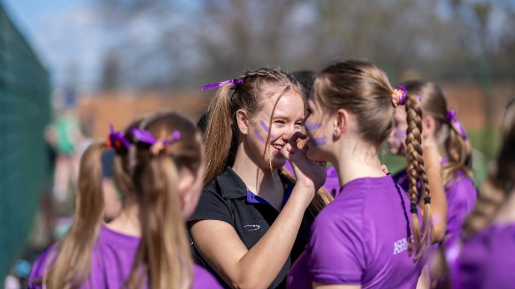 A young woman paints purple stripes on a teammate’s cheek whilst a group of Cheltenham College Houses athletes wearing purple shirts and ribbons gather outdoors.