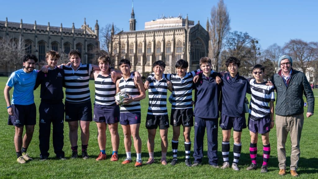 A Cheltenham College Houses rugby team and their coach standing in a line with arms round each other on a grassy pitch in front of a stone building.