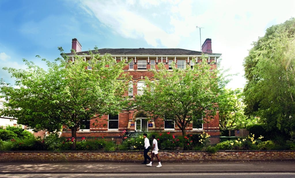 Two people walk along the pavement in front of a multi-storey red brick building framed by two large trees.