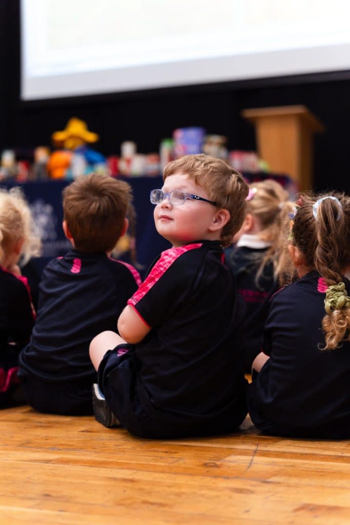 Students in black and pink uniforms sit on a wooden floor at a Cheltenham Prep Harvest Festival Assembly, with one boy in glasses looking towards the front of the room.