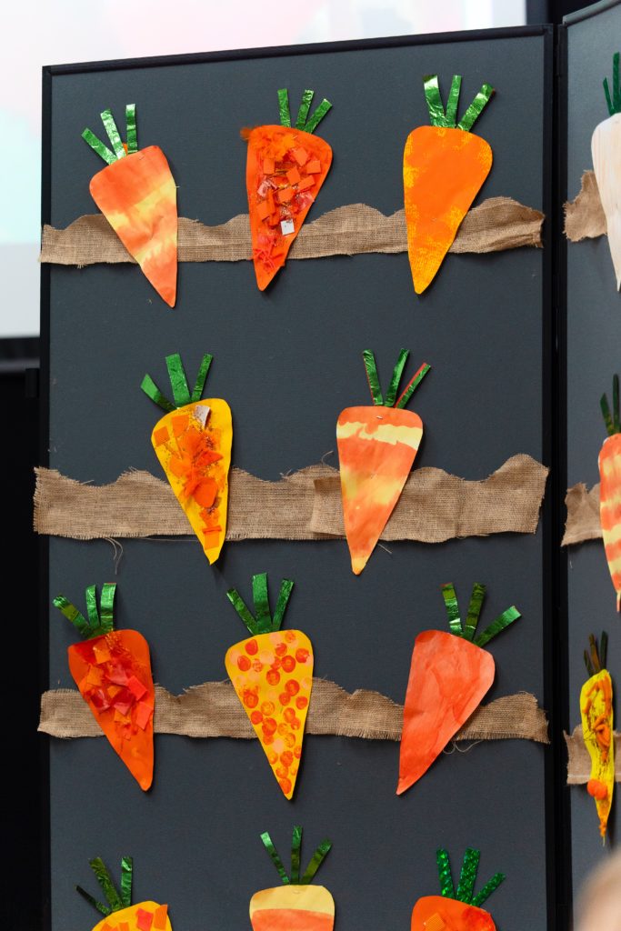 Paper carrot crafts with green foil tops attached to horizontal strips of hessian on a dark display board for the Cheltenham Prep Harvest Festival Assembly.