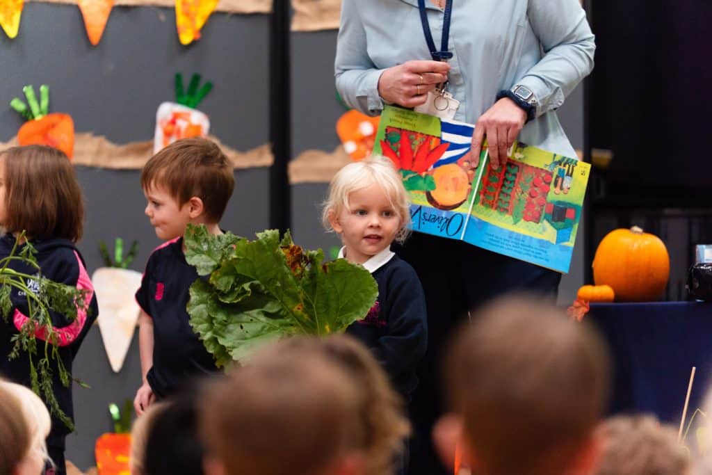 A young child holds a large leafy vegetable at a Cheltenham Prep Harvest Festival Assembly whilst an adult stands behind them holding an open picture book.