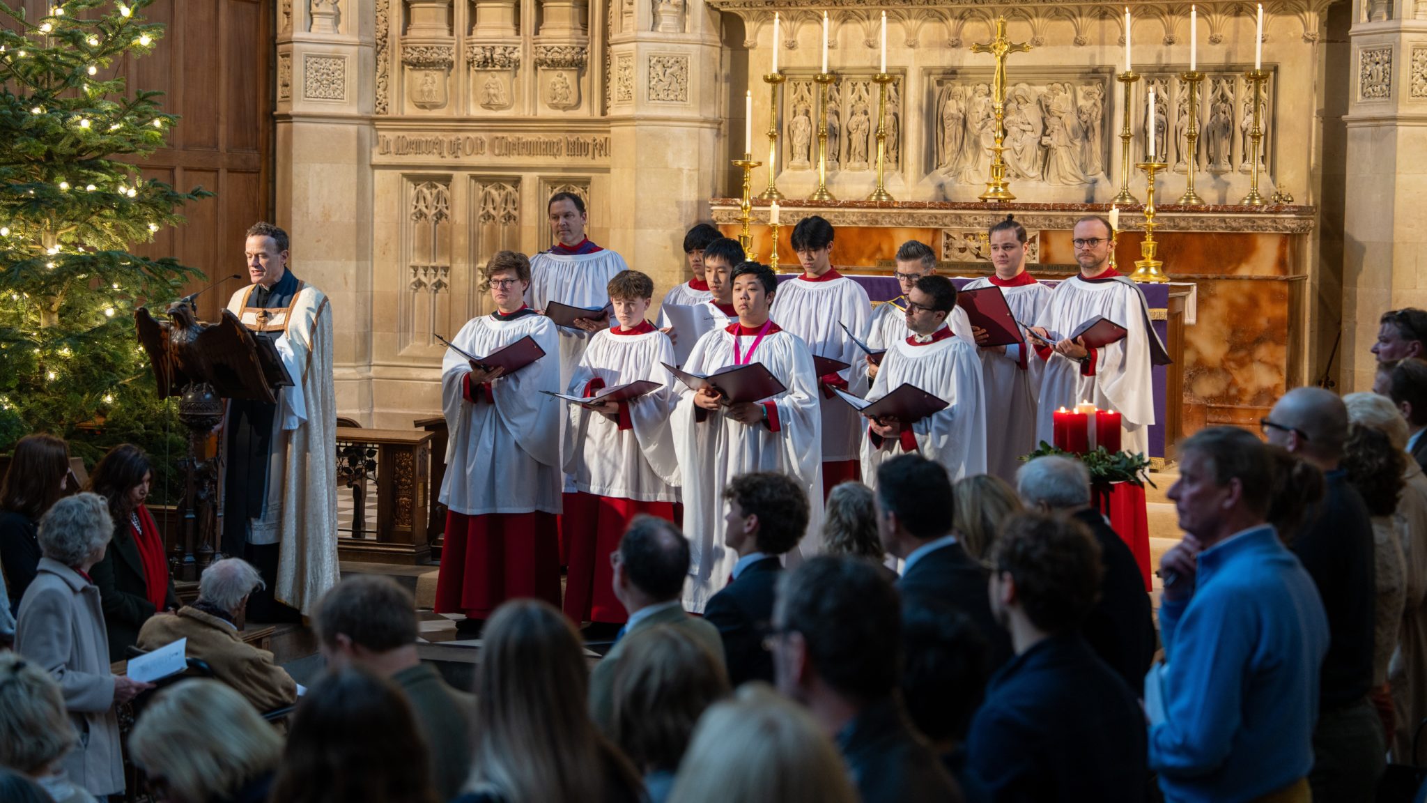 A robed choir sings from sheet music in a stone church, with an officiant standing at a lectern beside a large, lit Christmas tree.