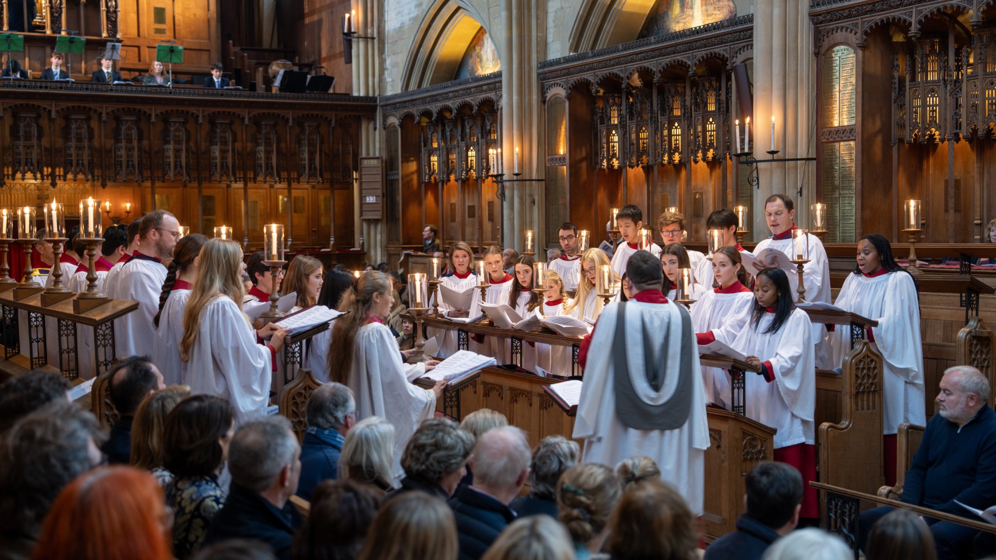 A choir in white robes stands in a church interior, holding sheet music and singing during a performance.