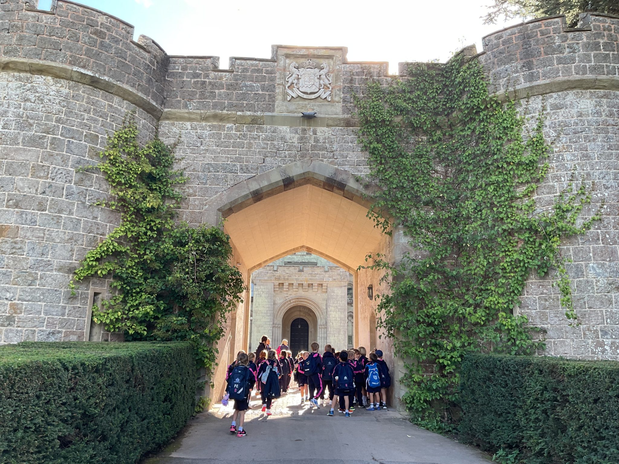 A group of children in school uniforms walk through a large stone archway towards a castle building.