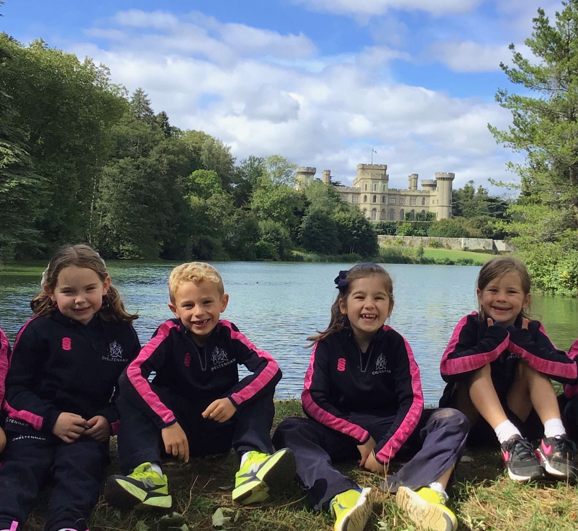 Four children in school tracksuits sit on the grass by a lake with a castle in the background.