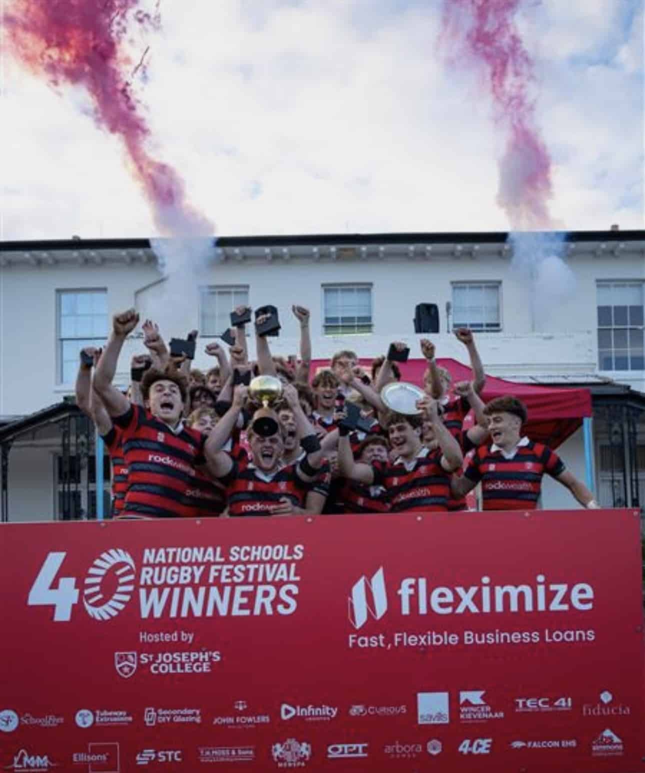 A rugby team in red and black striped jerseys celebrates on a stage, holding trophies aloft with pink smoke flares overhead, behind a banner identifying them as National Schools Rugby Festival winners.