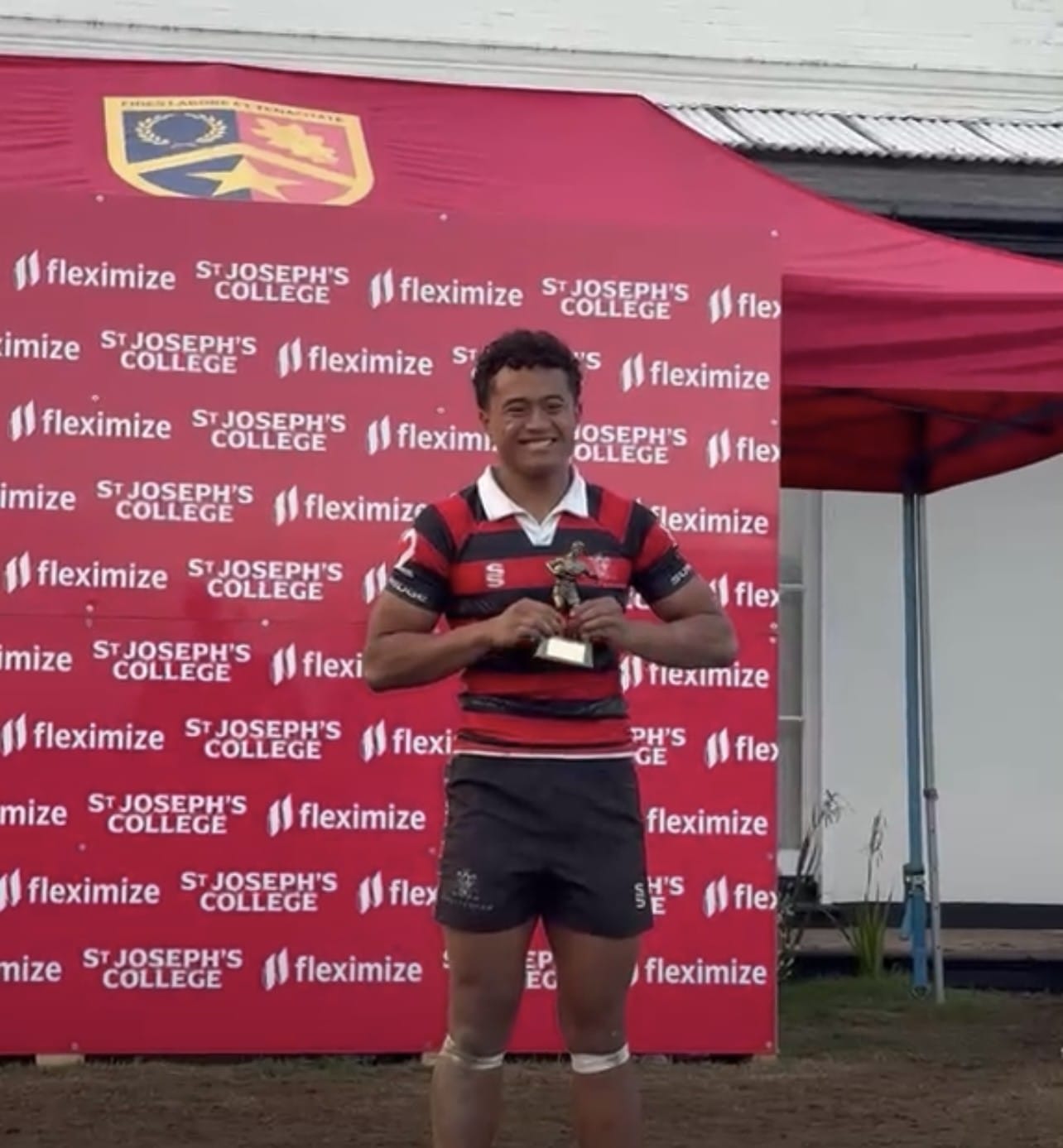 A smiling rugby player in a red and black striped jersey stands in front of a branded backdrop holding a trophy.