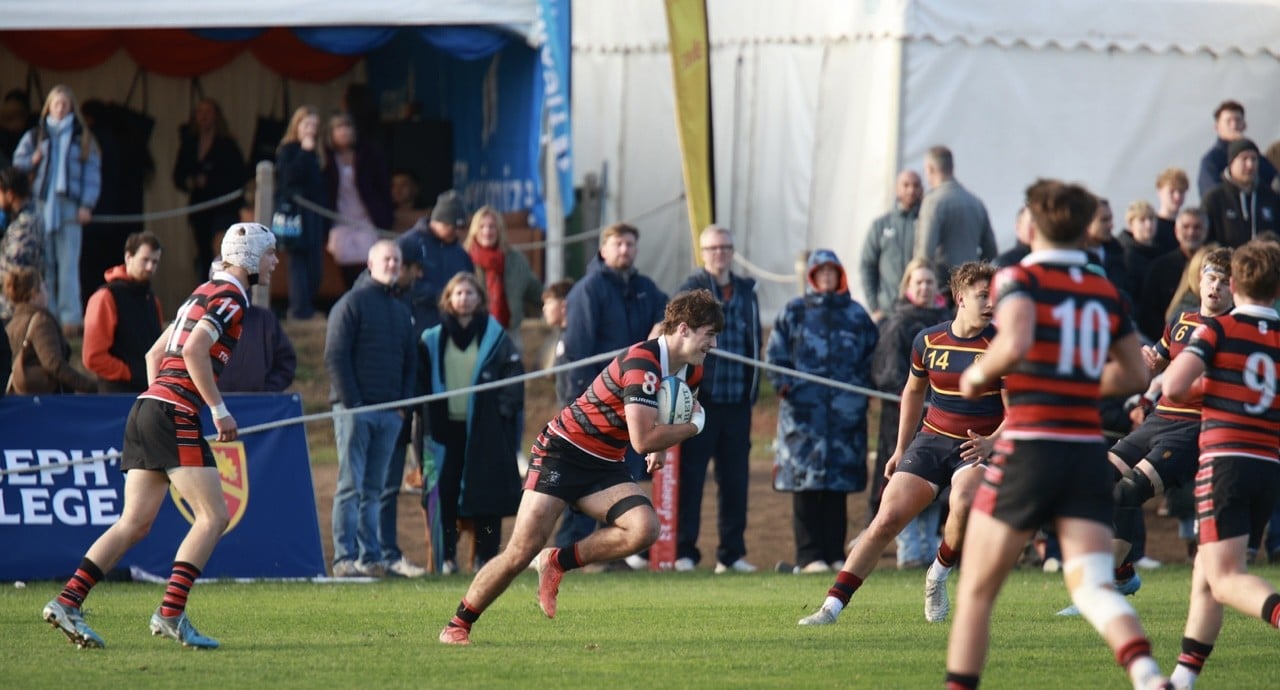 A rugby player in a red and black striped jersey runs with the ball on a grassy pitch during a match, whilst teammates and spectators watch from the touchline.
