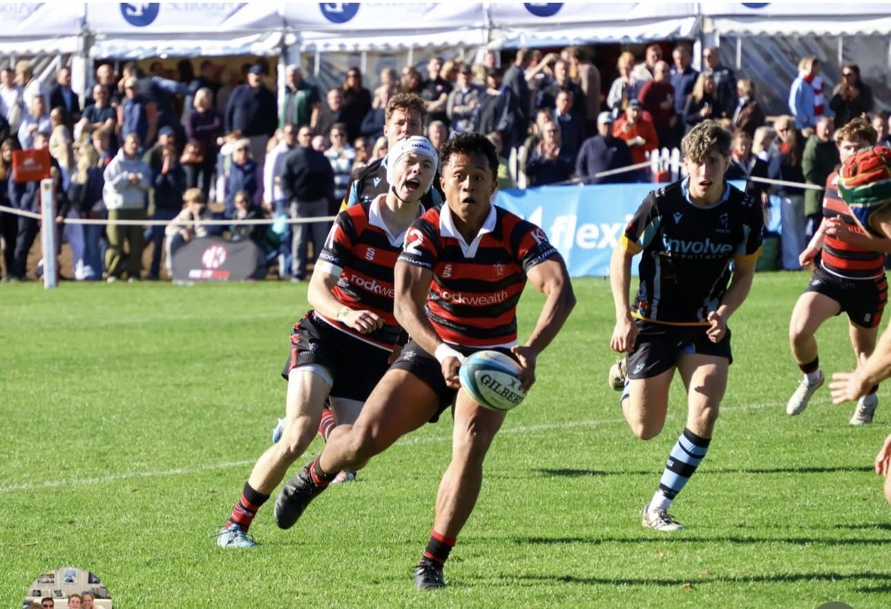 A rugby player in a red and black striped jumper runs with the ball whilst being chased by two opponents on a grass pitch.