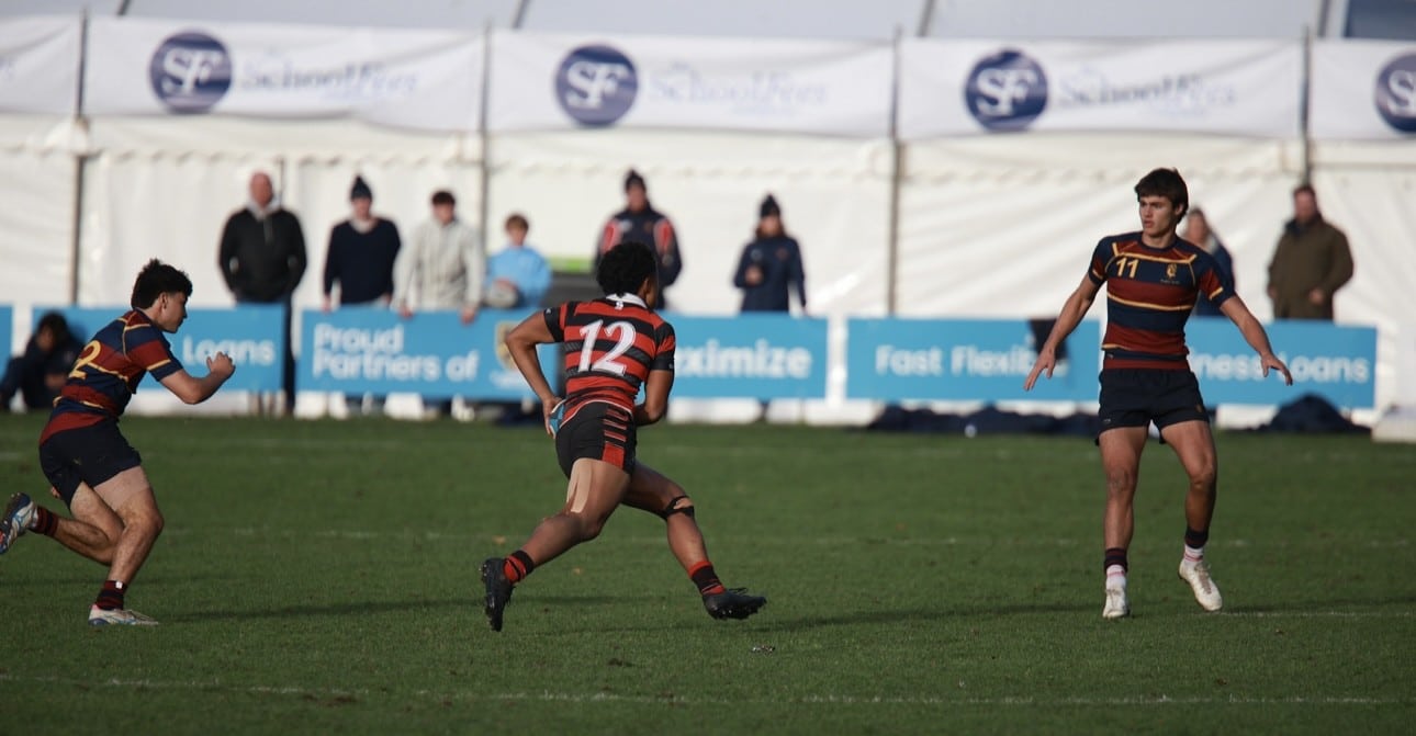 Three rugby players in maroon and gold jerseys on a pitch during a match.